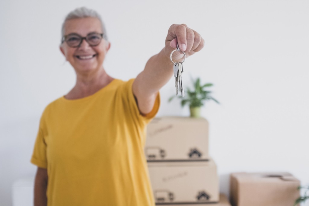 Senior happy woman holding keys of the new apartment standing in the empty home with moving boxes on the floor - concept of active elderly retired enjoying new life