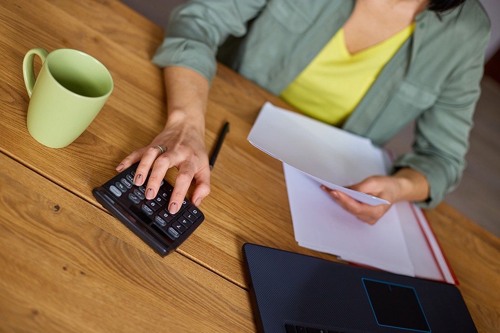 Close up of woman sit calculate business expenses on calculator at wooden table, modern workplace, freelancer working at home. Female busy manage family budget account expenditures.