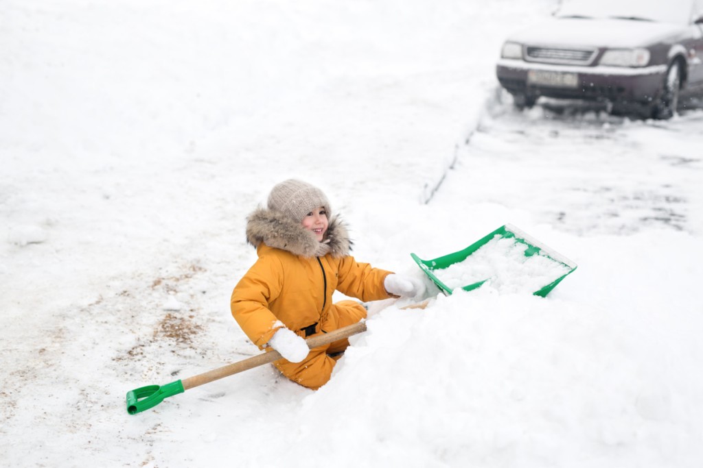 Little five-year-old girl in orange winter jumpsuit cleans snow big shovel. Snow removal after heavy snowfall. the child fell into a snowdrift of snow and laughs merrily