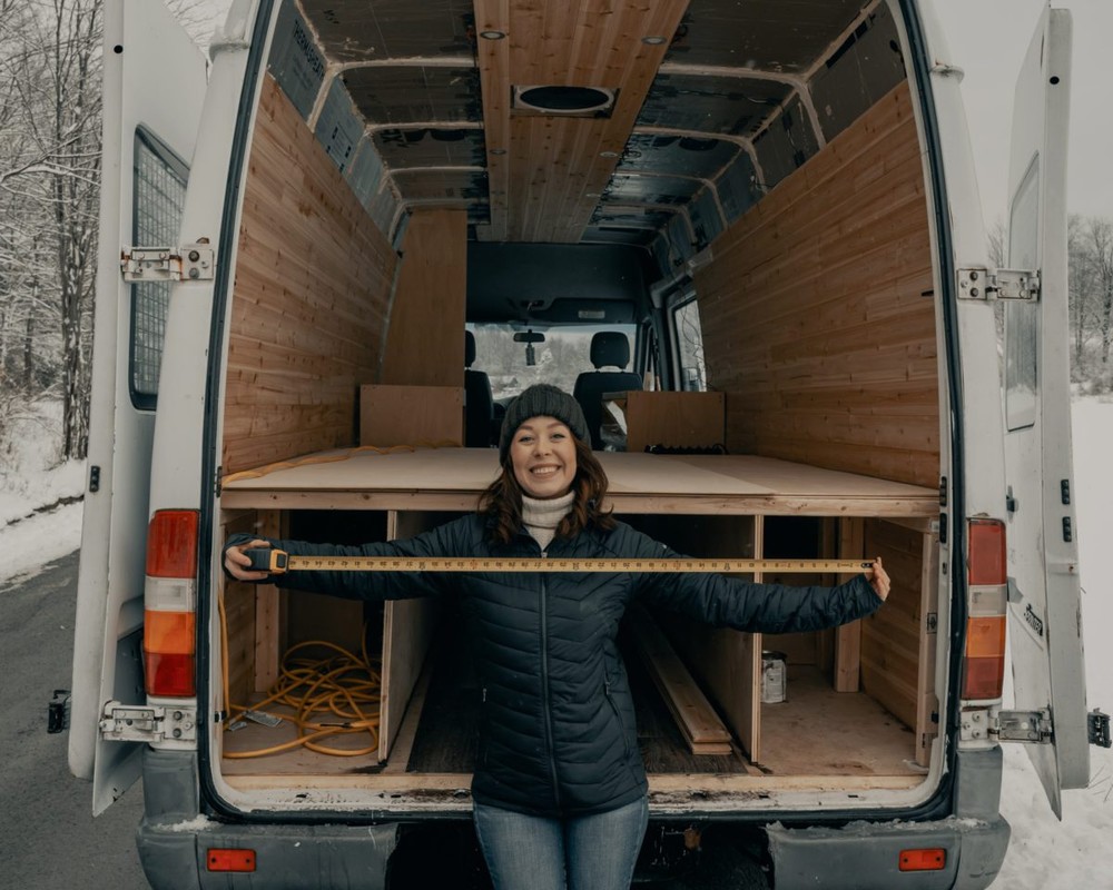 Millenial woman with a stretched out tape measure standing at the back of a removals van. The van doors are open and the van is empty