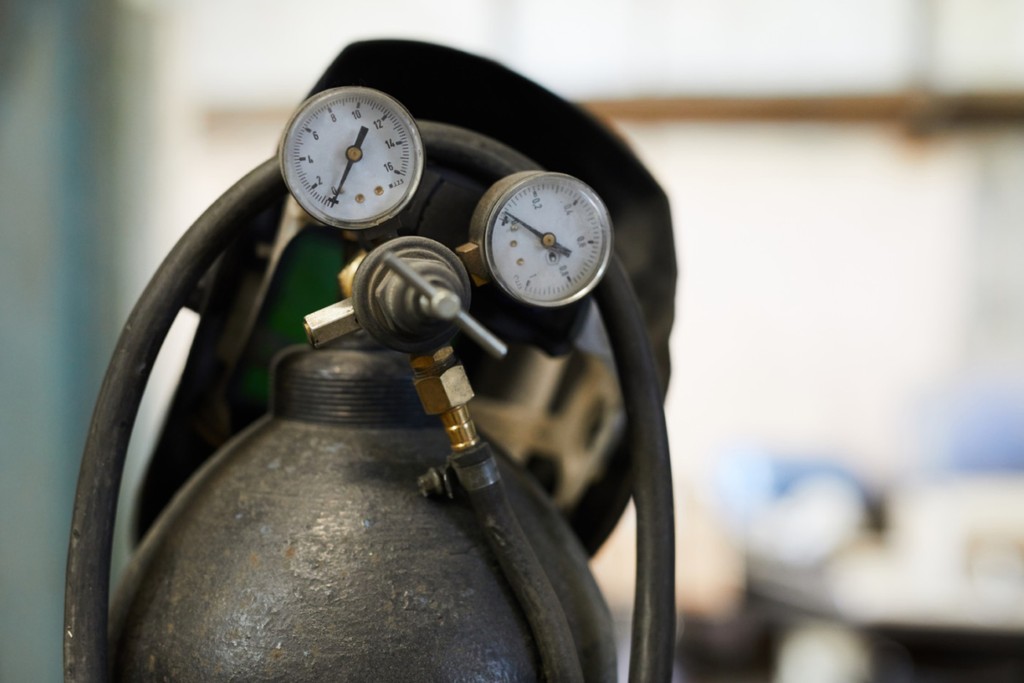 Close-up of air tank cylinder with tubes and pressure gauges, welding helmet hanging on gas cylinder