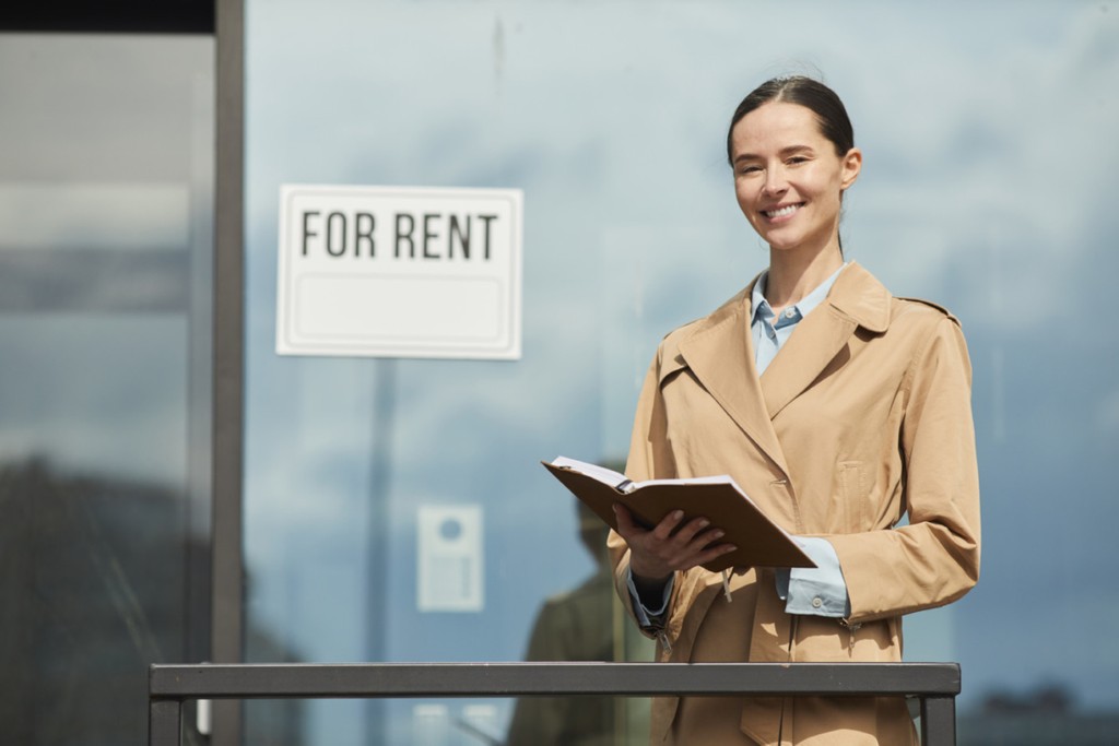 Waist up portrait of female real estate agent smiling happily looking at camera while standing next to FOR RENT sign outdoors